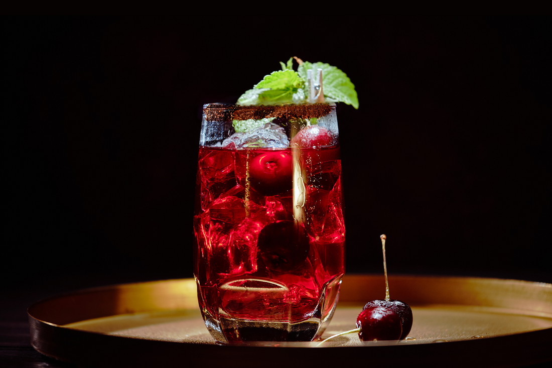Glass of a red spritzer drink on a gold tray, green garnish, cherries in a dark room
