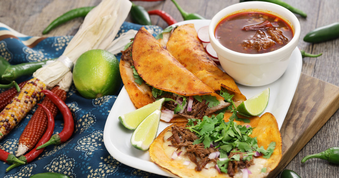 Birria Tacos on a plate with a small bowl of Birria. Corn, lime and chilis off to the side