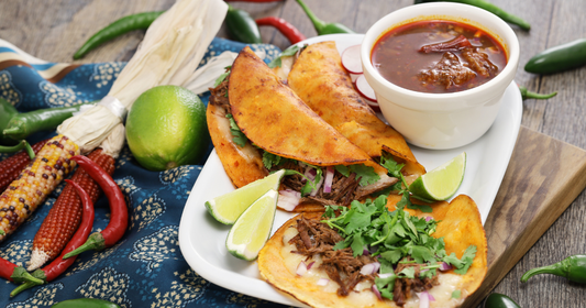 Birria Tacos on a plate with a small bowl of Birria. Corn, lime and chilis off to the side