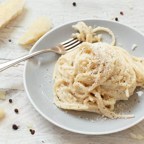 Plate of creamy pasta with parmesan and pepper on a white counter top. Whole peppercorns across the counter. 